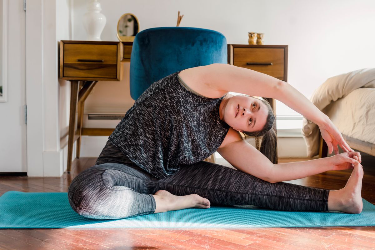 Woman doing a seated side stretch on a yoga mat at home, showing the ribcage and bra line area