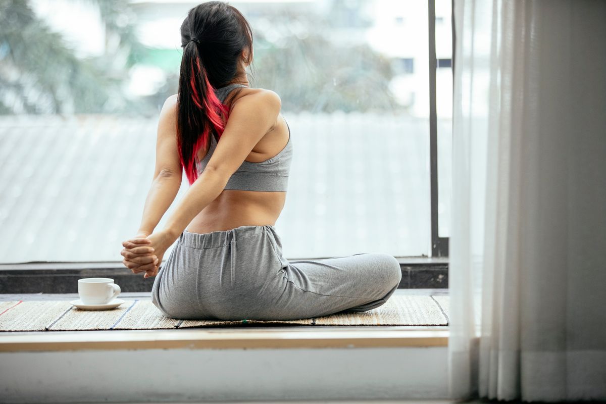 Woman seated on a mat by a window, reaching one arm behind her back to stretch the mid-back area